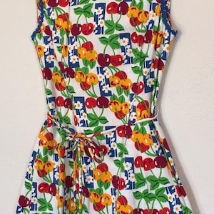 K&ouml;nnte beinhalten: &Auml;rmelloses wei&szlig;es Kleid mit einem lebendigen Kirschdruck in Rot, Gelb und Gr&uuml;n. Das Kleid hat eine blaue Bord&uuml;re am Ausschnitt und an den Armausschnitten sowie einen passenden Bindeg&uuml;rtel. Das Kleid h&auml;ngt an einem schwarzen Kleiderb&uuml;gel.