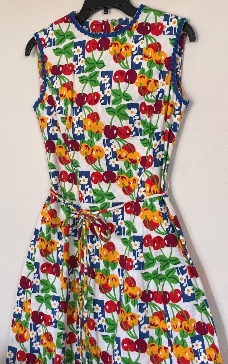 K&ouml;nnte beinhalten: &Auml;rmelloses wei&szlig;es Kleid mit einem lebendigen Kirschdruck in Rot, Gelb und Gr&uuml;n. Das Kleid hat eine blaue Bord&uuml;re am Ausschnitt und an den Armausschnitten sowie einen passenden Bindeg&uuml;rtel. Das Kleid h&auml;ngt an einem schwarzen Kleiderb&uuml;gel.