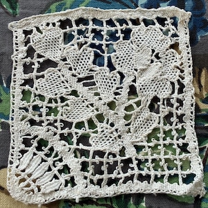 May include: A square piece of off-white needle lace featuring an intricate floral and leaf pattern. The design consists of interconnected heart-shaped petals and foliage motifs set against a delicate, open-work grid background with a finished border.