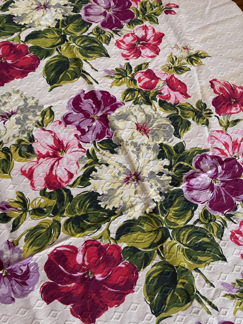 K&ouml;nnte beinhalten: Eine Tischdecke mit floralem Muster mit einem sich wiederholenden Muster aus gro&szlig;en, farbenfrohen Blumen. Die Blumen sind in Rot-, Rosa-, Lila- und Wei&szlig;t&ouml;nen gehalten, mit gr&uuml;nen Bl&auml;ttern. Der Hintergrund ist cremefarben mit einem dezenten Rautenmuster.