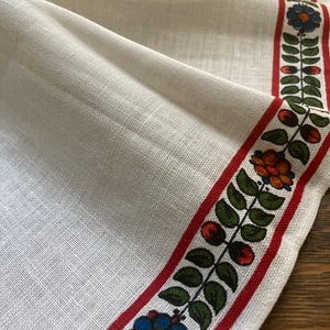 May include: Cream-colored linen table runner with a floral and leaf border. The border features red, green, blue, and orange flowers and leaves. The runner has a detailed floral design in the upper corner.