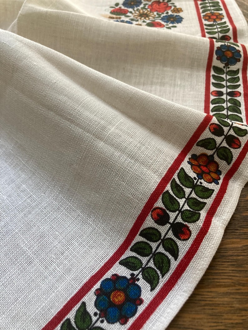 May include: Cream-colored linen table runner with a floral and leaf border. The border features red, green, blue, and orange flowers and leaves. The runner has a detailed floral design in the upper corner.
