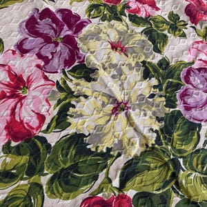 K&ouml;nnte beinhalten: Nahaufnahme eines floralen Stoffes mit gro&szlig;en, strukturierten Bl&uuml;ten in Rosa, Lila und Gelb. Die Blumen sind von gr&uuml;nen Bl&auml;ttern umgeben, wodurch ein lebendiges und detailliertes Muster entsteht. Der Hintergrund ist hellbeige.