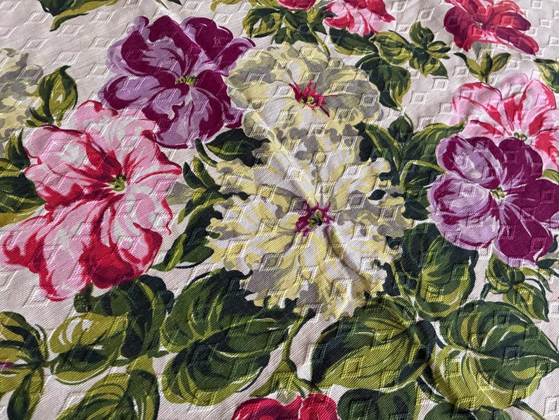 K&ouml;nnte beinhalten: Nahaufnahme eines floralen Stoffes mit gro&szlig;en, strukturierten Bl&uuml;ten in Rosa, Lila und Gelb. Die Blumen sind von gr&uuml;nen Bl&auml;ttern umgeben, wodurch ein lebendiges und detailliertes Muster entsteht. Der Hintergrund ist hellbeige.