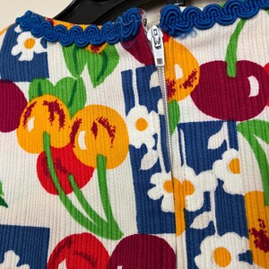 K&ouml;nnte beinhalten: Nahaufnahme eines wei&szlig;en Stoffes mit einem farbenfrohen Kirschmuster. Das Design zeigt rote, gelbe und violette Kirschen mit gr&uuml;nen Stielen und Bl&auml;ttern, vor einem blau-wei&szlig;en Hintergrund. Eine blaue Bord&uuml;re und ein Rei&szlig;verschluss sind sichtbar.