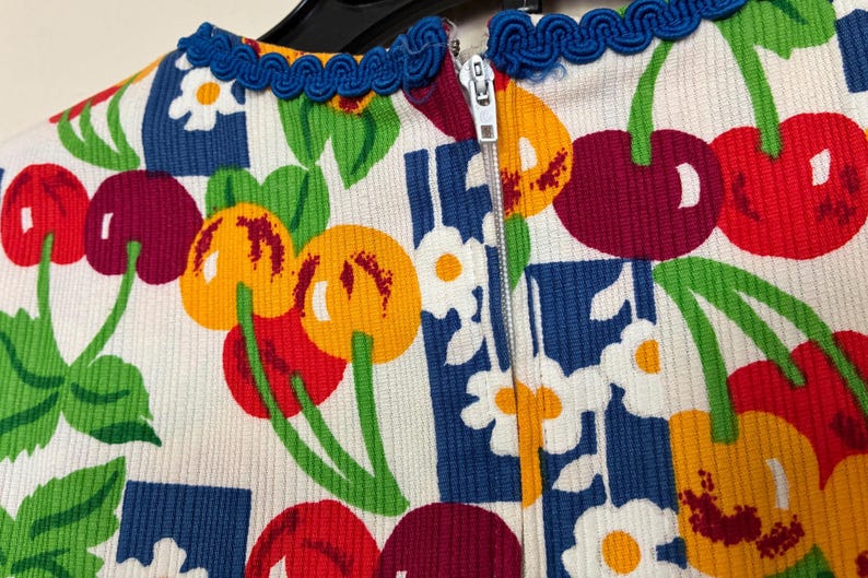 K&ouml;nnte beinhalten: Nahaufnahme eines wei&szlig;en Stoffes mit einem farbenfrohen Kirschmuster. Das Design zeigt rote, gelbe und violette Kirschen mit gr&uuml;nen Stielen und Bl&auml;ttern, vor einem blau-wei&szlig;en Hintergrund. Eine blaue Bord&uuml;re und ein Rei&szlig;verschluss sind sichtbar.