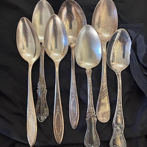 May include: A collection of seven silver-toned metal spoons with varied handle designs, including plain oval accents, intricate floral engravings, and decorative scrollwork patterns, arranged together on a dark fabric surface.