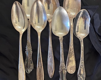 Könnte beinhalten: Eine Sammlung von sieben silberfarbenen Metalllöffeln mit unterschiedlichen Griffdesigns, darunter schlichte ovale Akzente, komplexe florale Gravuren und dekorative Schnörkelmuster, angeordnet auf einem dunklen Stoff.