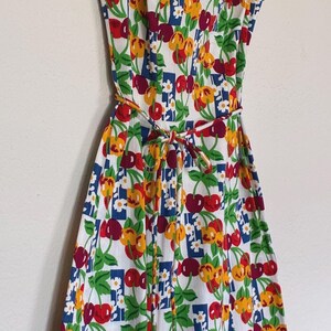 K&ouml;nnte beinhalten: &Auml;rmelloses wei&szlig;es Kleid mit einem lebendigen Kirschdruck in Rot, Gelb und Gr&uuml;n. Das Kleid hat eine blaue Bord&uuml;re und einen passenden Bindeg&uuml;rtel. Ein lustiges Sommerkleid im Retro-Stil.