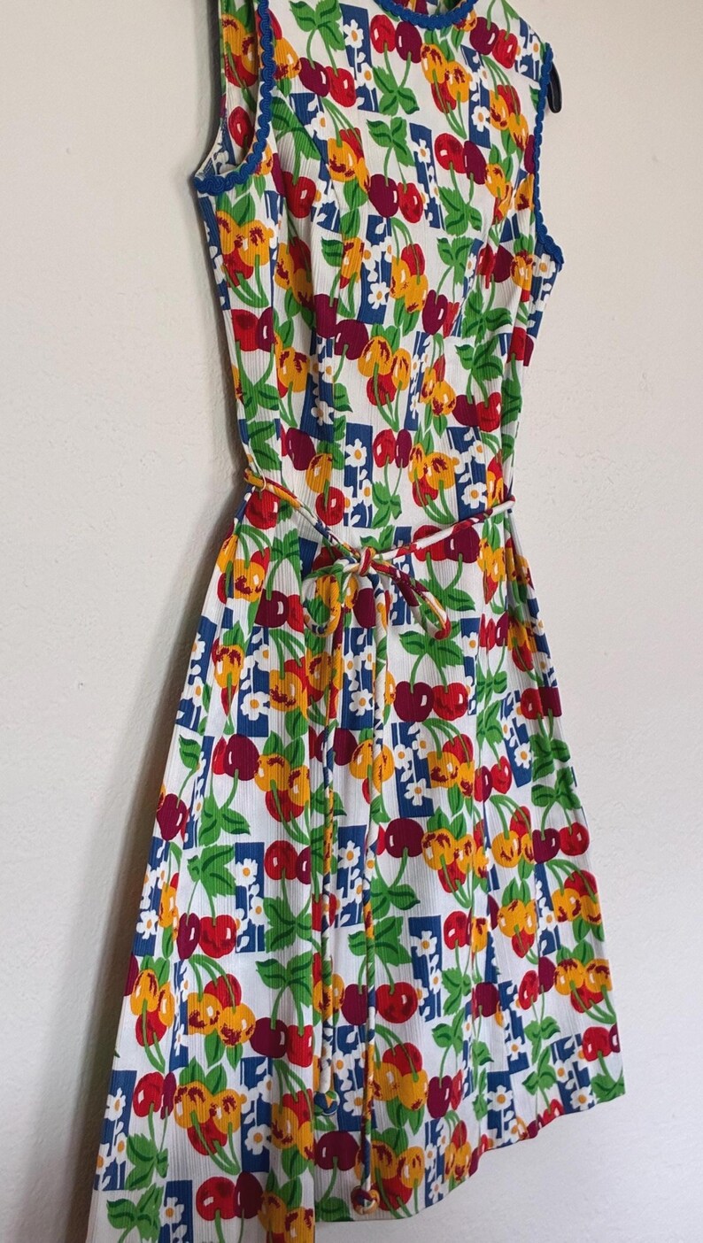K&ouml;nnte beinhalten: &Auml;rmelloses wei&szlig;es Kleid mit einem lebendigen Kirschdruck in Rot, Gelb und Gr&uuml;n. Das Kleid hat eine blaue Bord&uuml;re und einen passenden Bindeg&uuml;rtel. Ein lustiges Sommerkleid im Retro-Stil.