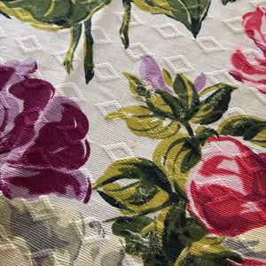 K&ouml;nnte beinhalten: Nahaufnahme eines floralen Stoffes mit Rautenmuster. Das Design zeigt gro&szlig;e Rosen in Rot- und Lilat&ouml;nen mit gr&uuml;nen und gelben Bl&auml;ttern. Der Hintergrund ist cremefarben.