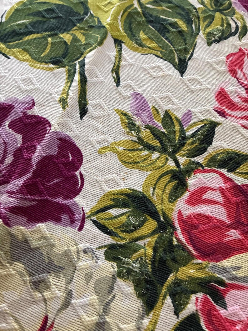 K&ouml;nnte beinhalten: Nahaufnahme eines floralen Stoffes mit Rautenmuster. Das Design zeigt gro&szlig;e Rosen in Rot- und Lilat&ouml;nen mit gr&uuml;nen und gelben Bl&auml;ttern. Der Hintergrund ist cremefarben.
