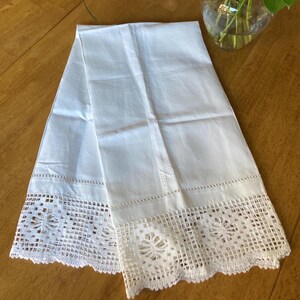 May include: White linen towel with a decorative crocheted lace border. The towel has an embroidered initial and a delicate stitched edge. The lace features a floral design. The towel is folded on a wooden surface.