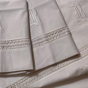May include: White bed linens with embroidered capital letter "R" and decorative cutwork along the edges. The linens appear to be made of a soft, high-quality fabric, suggesting a luxurious bedding set.