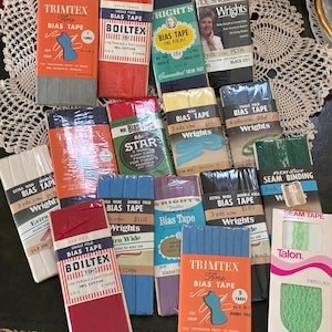 May include: A collection of vintage bias tape packages in various colors, including red, blue, green, and gray. The packages feature brands like Trimtex, Wrights, and Boiltex, with text indicating yardage and material. The tapes are for sewing and decorative trimming.
