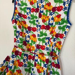 K&ouml;nnte beinhalten: &Auml;rmelloses Kleid mit einem lebendigen Kirschmuster in Rot, Gelb und Burgunderrot auf wei&szlig;em Grund. Das Kleid hat eine blaue Bord&uuml;re am Ausschnitt und an den Armausschnitten sowie einen passenden G&uuml;rtel. Der Stoff hat eine strukturierte Cord-Optik.