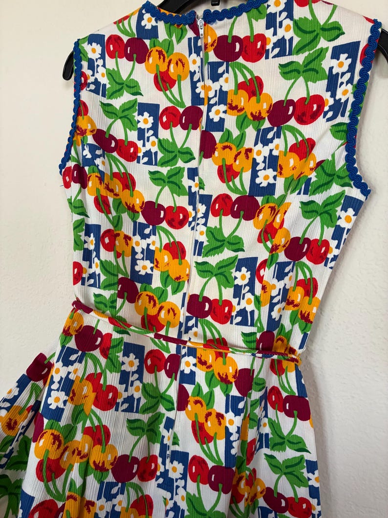K&ouml;nnte beinhalten: &Auml;rmelloses Kleid mit einem lebendigen Kirschmuster in Rot, Gelb und Burgunderrot auf wei&szlig;em Grund. Das Kleid hat eine blaue Bord&uuml;re am Ausschnitt und an den Armausschnitten sowie einen passenden G&uuml;rtel. Der Stoff hat eine strukturierte Cord-Optik.
