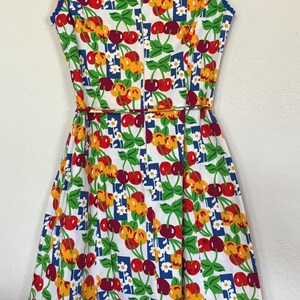 K&ouml;nnte beinhalten: Ein &auml;rmelloses Kleid im Vintage-Stil mit einem lebendigen Kirschmuster. Der wei&szlig;e Stoff ist mit roten, gelben und gr&uuml;nen Kirschen verziert, die durch blaue und wei&szlig;e Blumenquadrate akzentuiert werden. Das Kleid hat ein figurbetontes Oberteil, eine betonte Taille und einen ausgestellten Rock.