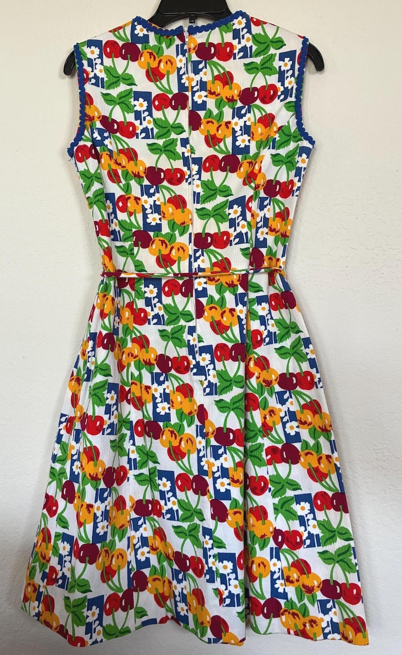 K&ouml;nnte beinhalten: Ein &auml;rmelloses Kleid im Vintage-Stil mit einem lebendigen Kirschmuster. Der wei&szlig;e Stoff ist mit roten, gelben und gr&uuml;nen Kirschen verziert, die durch blaue und wei&szlig;e Blumenquadrate akzentuiert werden. Das Kleid hat ein figurbetontes Oberteil, eine betonte Taille und einen ausgestellten Rock.