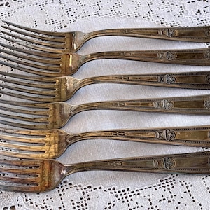 May include: Six antique silver forks with ornate detailing on the handles. The forks are arranged on a white lace tablecloth. The tines of the forks are visible, and the silver has a slightly tarnished appearance.