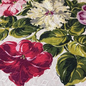 K&ouml;nnte beinhalten: Nahaufnahme eines floralen Stoffes mit gro&szlig;en, farbenfrohen Blumen in Rot-, Rosa-, Lila- und Cremet&ouml;nen. Die Blumen sind von gr&uuml;nen Bl&auml;ttern auf einem wei&szlig;en Hintergrund mit einem dezenten Rautenmuster umgeben.
