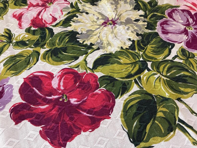 K&ouml;nnte beinhalten: Nahaufnahme eines floralen Stoffes mit gro&szlig;en, farbenfrohen Blumen in Rot-, Rosa-, Lila- und Cremet&ouml;nen. Die Blumen sind von gr&uuml;nen Bl&auml;ttern auf einem wei&szlig;en Hintergrund mit einem dezenten Rautenmuster umgeben.