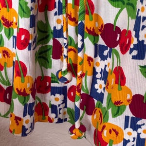 K&ouml;nnte beinhalten: Nahaufnahme eines wei&szlig;en Stoffes mit einem lebendigen Kirschmuster. Das Design zeigt rote, gelbe und gr&uuml;ne Kirschen sowie wei&szlig;e Blumen und blaue geometrische Formen. Der Stoff hat eine gerippte Textur und ein Kordelzugdetail.