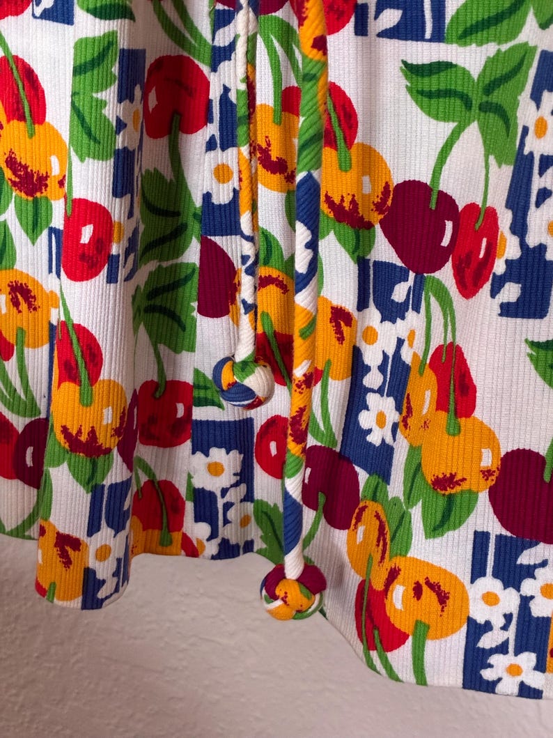 K&ouml;nnte beinhalten: Nahaufnahme eines wei&szlig;en Stoffes mit einem lebendigen Kirschmuster. Das Design zeigt rote, gelbe und gr&uuml;ne Kirschen sowie wei&szlig;e Blumen und blaue geometrische Formen. Der Stoff hat eine gerippte Textur und ein Kordelzugdetail.