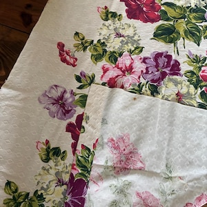 K&ouml;nnte beinhalten: Cremefarbener Stoff mit floralem Muster und sich wiederholendem Rautenmuster. Der Stoff ist mit gro&szlig;en, leuchtenden Blumen in Rot-, Rosa-, Lila- und Wei&szlig;t&ouml;nen mit gr&uuml;nem Laub verziert. Der Stoff wird wahrscheinlich f&uuml;r Heimdekoration oder Bastelarbeiten verwendet.