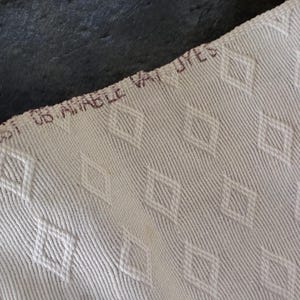 K&ouml;nnte beinhalten: Nahaufnahme eines cremefarbenen Stoffes mit Rautenmuster. Der Stoff hat eine strukturierte Oberfl&auml;che mit diagonalen Linien. Der Text "BCSI US AMABLE VAT DYES" ist braun am oberen Rand des Stoffes aufgedruckt.