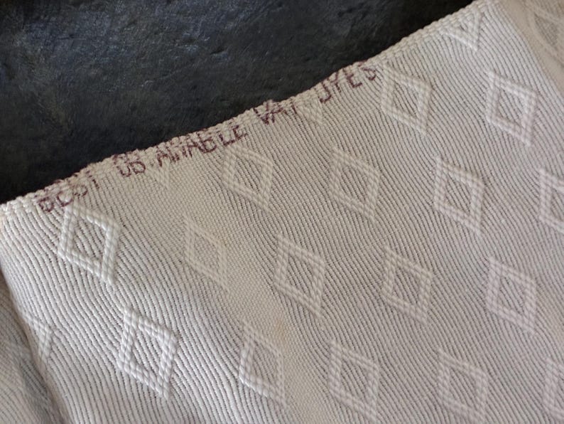 K&ouml;nnte beinhalten: Nahaufnahme eines cremefarbenen Stoffes mit Rautenmuster. Der Stoff hat eine strukturierte Oberfl&auml;che mit diagonalen Linien. Der Text "BCSI US AMABLE VAT DYES" ist braun am oberen Rand des Stoffes aufgedruckt.