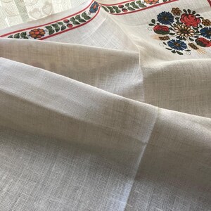 May include: Off-white linen tablecloth with a floral design. The border features a red stripe with green leaves and red berries. The center has a floral bouquet with red, blue, and yellow flowers.