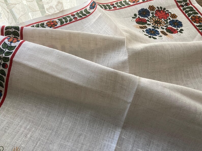 May include: Off-white linen tablecloth with a floral design. The border features a red stripe with green leaves and red berries. The center has a floral bouquet with red, blue, and yellow flowers.