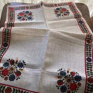 May include: A white linen tablecloth with a floral design. The tablecloth features a red border with green leaves and colorful flowers. The center of the tablecloth has four floral bouquets with red, blue, and yellow flowers.