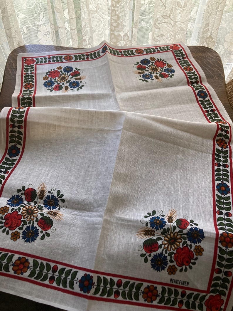 May include: A white linen tablecloth with a floral design. The tablecloth features a red border with green leaves and colorful flowers. The center of the tablecloth has four floral bouquets with red, blue, and yellow flowers.