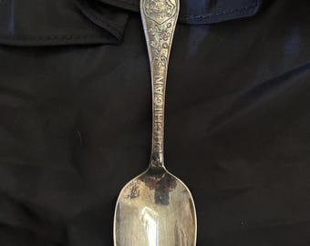 Könnte beinhalten: Ein silberfarbener Souvenir-Löffel mit einem geprägten Adler und Schild am oberen Ende des Griffs, einem Wappen darunter und dem vertikal eingravierten Wort MICHIGAN entlang des Stiels, das in eine glatte, ovale Löffelschale übergeht.