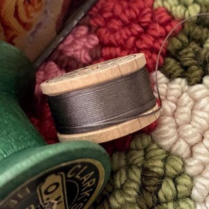 May include: A wooden spool of dark gray thread positioned next to a larger green wooden spool with a black label reading Clark's O.N.T. The spools rest against a textured background featuring a pattern of red, green, and cream-colored loops.