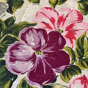 K&ouml;nnte beinhalten: Nahaufnahme eines Blumenmusters mit gro&szlig;en, stilisierten Bl&uuml;ten in Rosa-, Rot- und Lilat&ouml;nen. Die Blumen sind vor einem cremefarbenen Hintergrund mit einem dezenten Rautenmuster angeordnet, umgeben von gr&uuml;nen Bl&auml;ttern.