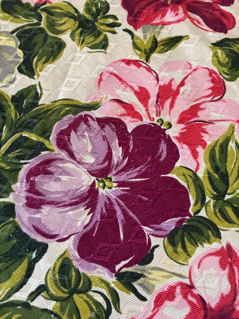 K&ouml;nnte beinhalten: Nahaufnahme eines Blumenmusters mit gro&szlig;en, stilisierten Bl&uuml;ten in Rosa-, Rot- und Lilat&ouml;nen. Die Blumen sind vor einem cremefarbenen Hintergrund mit einem dezenten Rautenmuster angeordnet, umgeben von gr&uuml;nen Bl&auml;ttern.