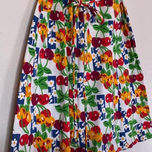 K&ouml;nnte beinhalten: Ein wei&szlig;es Kleid mit einem lebendigen Kirschmuster in Rot, Gelb und Gr&uuml;n. Das Design umfasst blaue Quadrate und kleine wei&szlig;e Blumen. Das Kleid hat eine Bindetailie in der Taille.