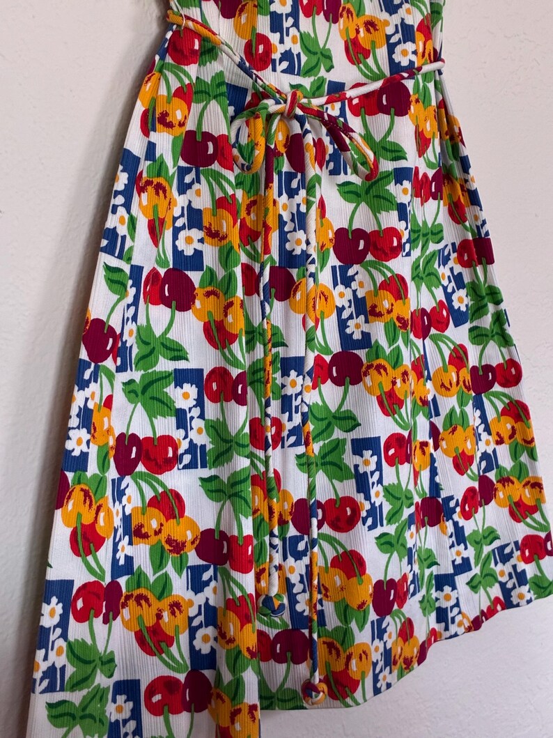 K&ouml;nnte beinhalten: Ein wei&szlig;es Kleid mit einem lebendigen Kirschmuster in Rot, Gelb und Gr&uuml;n. Das Design umfasst blaue Quadrate und kleine wei&szlig;e Blumen. Das Kleid hat eine Bindetailie in der Taille.