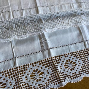 May include: Close-up of a white linen textile with intricate crochet and embroidered details. The design features floral patterns and scalloped edges, showcasing a vintage aesthetic. The fabric is folded, revealing the texture and craftsmanship.