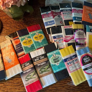 May include: A collection of vintage bias tape packages in various colors, including navy, orange, red, green, yellow, and blue. The packages feature brands like Wrights, Boiltex, and Talon, with text indicating the tape's length and type.