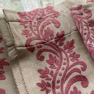 May include: A beige fabric curtain featuring a raised, textured pattern in a deep rose pink color. The design consists of intricate, swirling floral and leaf motifs that run vertically across the herringbone-weave base material of the textile.