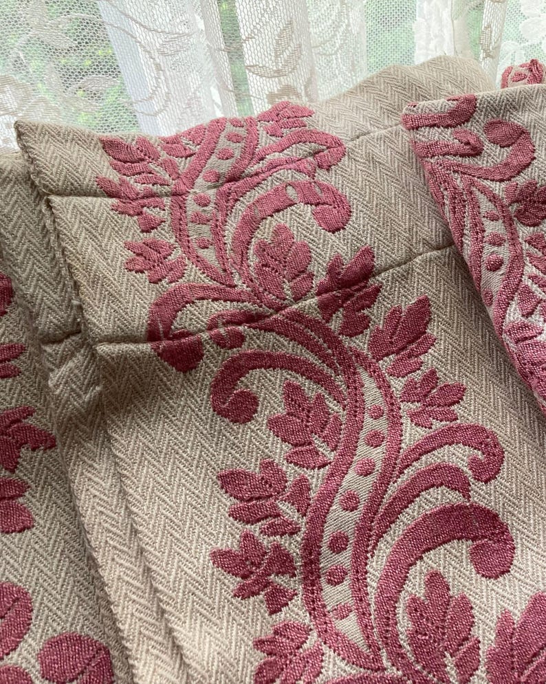 May include: A beige fabric curtain featuring a raised, textured pattern in a deep rose pink color. The design consists of intricate, swirling floral and leaf motifs that run vertically across the herringbone-weave base material of the textile.
