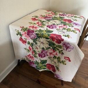 K&ouml;nnte beinhalten: Cremefarbene Tischdecke mit floralem Muster aus roten, rosa, lila und wei&szlig;en Blumen und gr&uuml;nen Bl&auml;ttern. Die Tischdecke bedeckt einen quadratischen Tisch, mit einem Holzstuhl auf der rechten Seite.