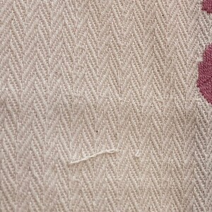 May include: A beige fabric with a woven herringbone pattern featuring a textured surface. On the right side, there is a section of dark pink embroidery displaying a curved, ornate floral design that contrasts with the neutral base fabric.