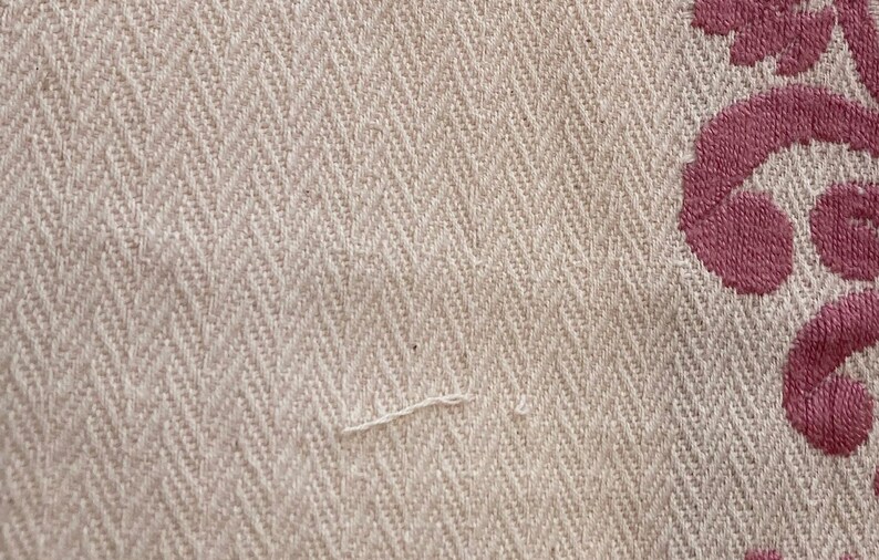 May include: A beige fabric with a woven herringbone pattern featuring a textured surface. On the right side, there is a section of dark pink embroidery displaying a curved, ornate floral design that contrasts with the neutral base fabric.