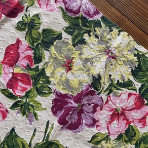 K&ouml;nnte beinhalten: Nahaufnahme eines floralen Stoffes mit gro&szlig;en, farbenfrohen Blumen in Rosa, Lila und Gelb vor einem wei&szlig;en Hintergrund mit Rautenmuster. Gr&uuml;ne Bl&auml;tter und Stiele setzen Kontraste. Der Stoff ist wahrscheinlich f&uuml;r Heimdekoration oder Kleidung gedacht.