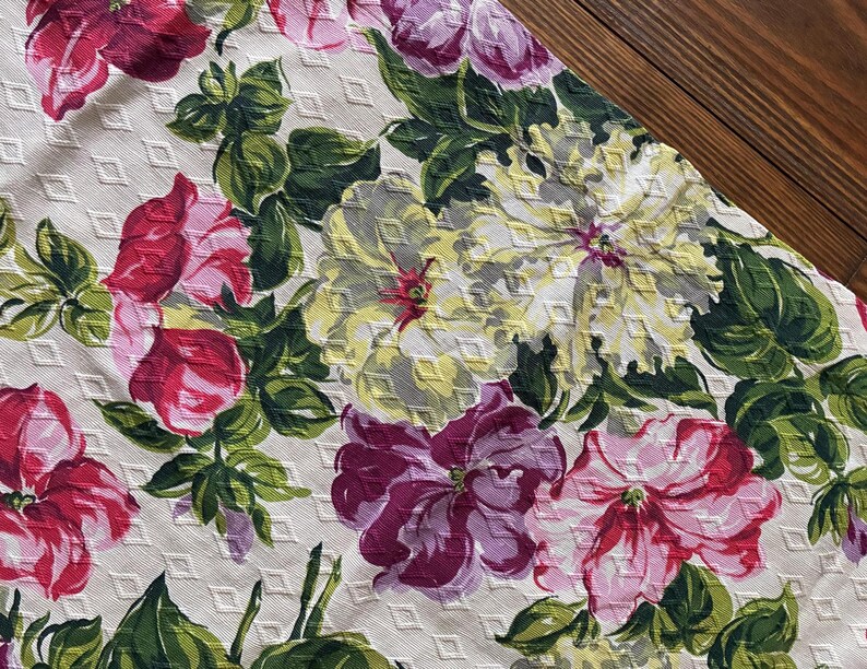 K&ouml;nnte beinhalten: Nahaufnahme eines floralen Stoffes mit gro&szlig;en, farbenfrohen Blumen in Rosa, Lila und Gelb vor einem wei&szlig;en Hintergrund mit Rautenmuster. Gr&uuml;ne Bl&auml;tter und Stiele setzen Kontraste. Der Stoff ist wahrscheinlich f&uuml;r Heimdekoration oder Kleidung gedacht.